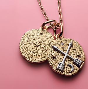 Pandora Talisman gold medallion necklace featuring crossed arrows, stars, and crescent moon design on textured gold discs, displayed against a soft pink background.