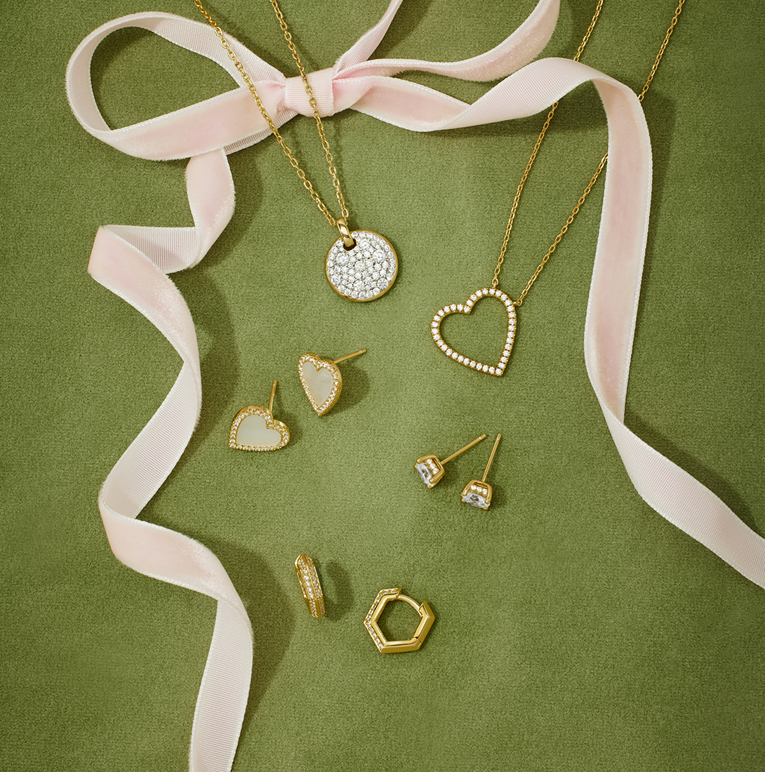 Ania Haie 14k gold-plated and cubic zirconia jewelry collection styled on a green velvet background with soft pink ribbon — featuring open heart and disc pendant necklaces, mother-of-pearl heart studs, crystal solitaire studs, and hexagon huggie hoops for a chic, modern look.