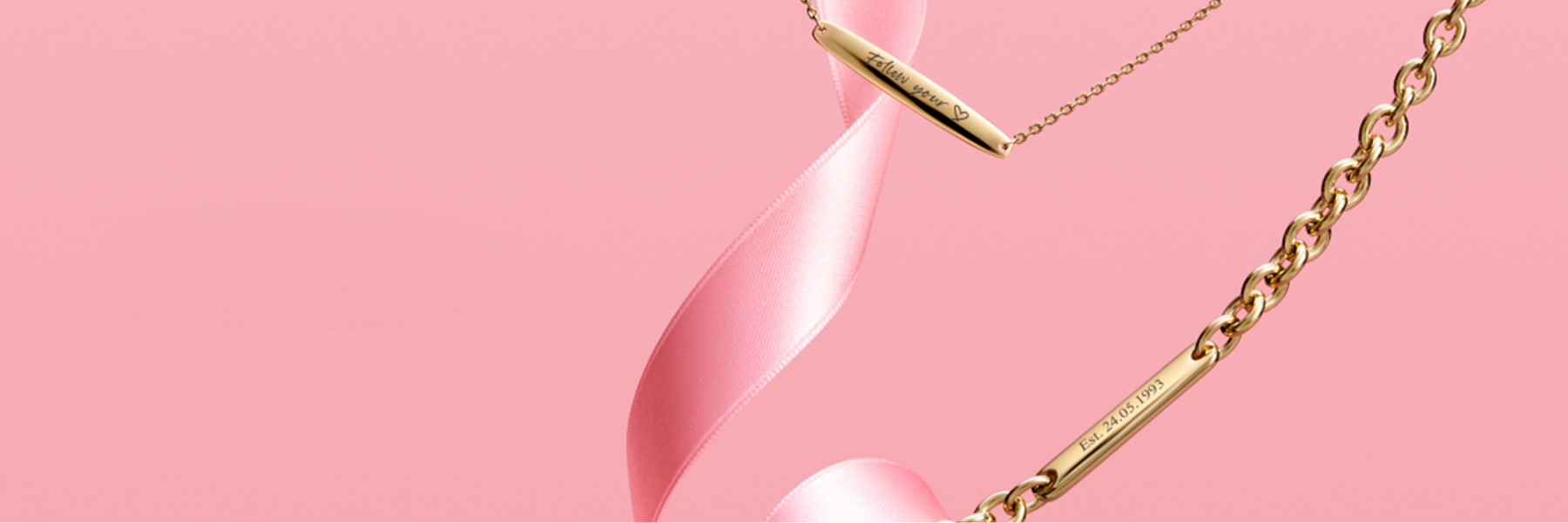Engravable jewelry featuring gold bar necklaces and bracelets with personalized inscriptions, styled with a pink ribbon on a soft pink background.