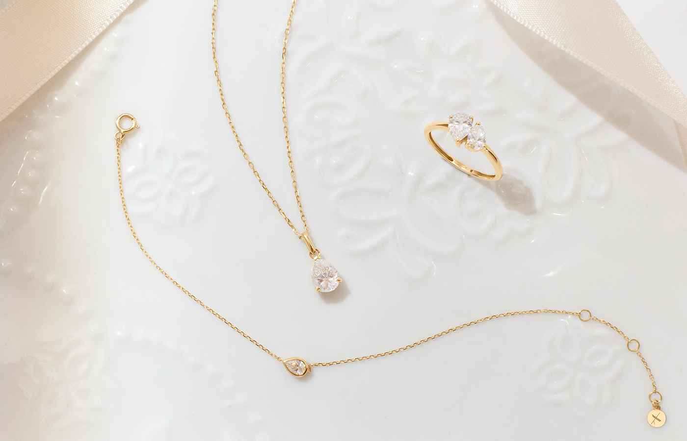 Aurelie Gi jewelry in 14k gold featuring a pear-shaped solitaire necklace, matching delicate bracelet, and a three-stone pear and round-cut ring, displayed on an elegant white embossed ceramic plate.