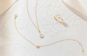 Aurelie Gi jewelry in 14k gold featuring a pear-shaped solitaire necklace, matching delicate bracelet, and a three-stone pear and round-cut ring, displayed on an elegant white embossed ceramic plate.