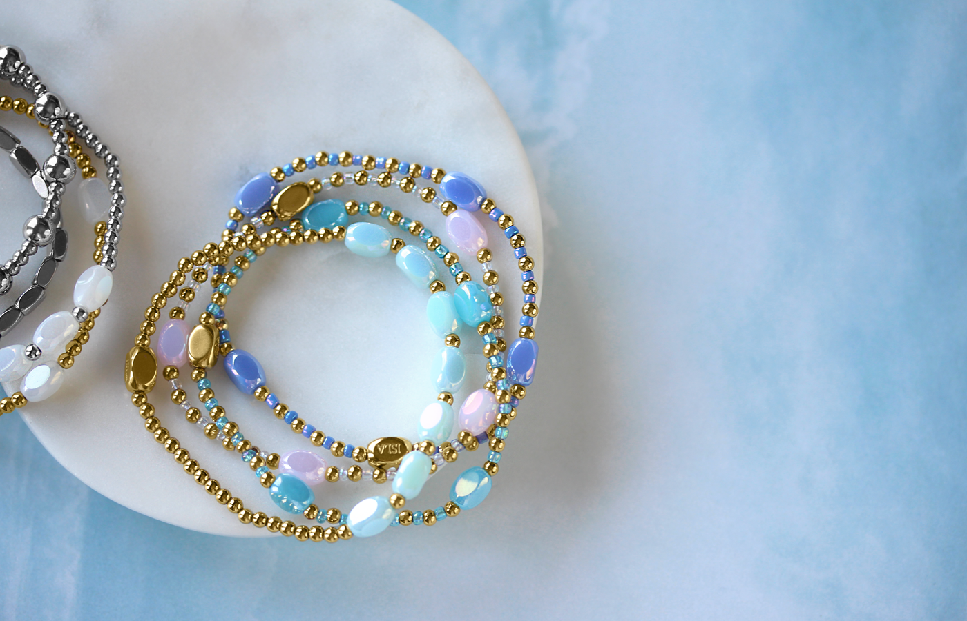 Layered stretchy waterproof bracelets featuring gold accents with stunning blue or pink beads, elegantly arranged on a white dish. Vibrant silver options with white beads complement the display, set against a soothing waterwash blue background.