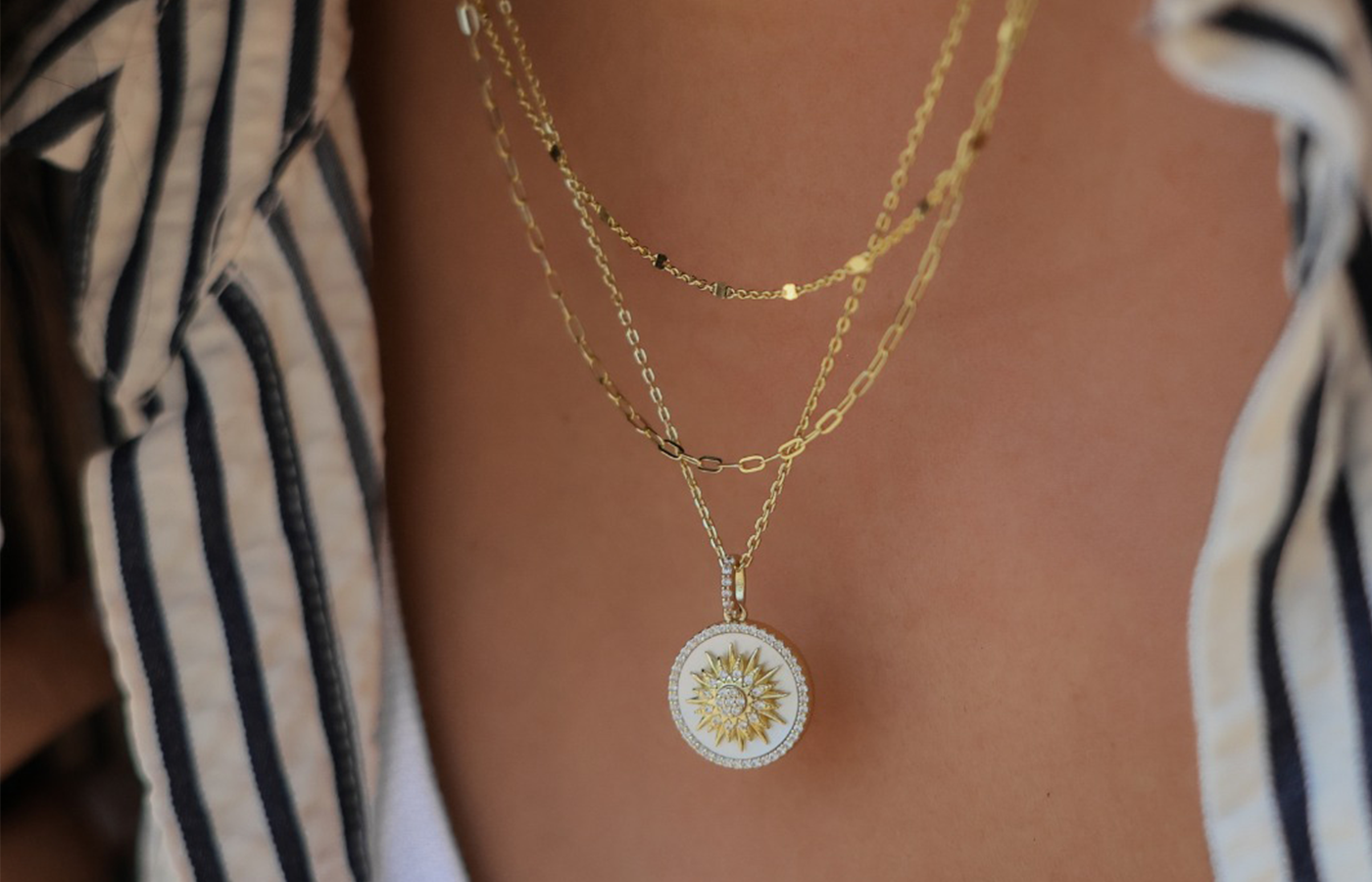 Lafonn jewelry layered gold chain necklaces featuring a round white enamel pendant with a gold sunburst and sparkling center stone, styled over a white tank top and striped shirt for a chic, modern look.