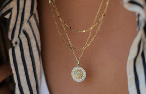 Lafonn jewelry layered gold chain necklaces featuring a round white enamel pendant with a gold sunburst and sparkling center stone, styled over a white tank top and striped shirt for a chic, modern look.