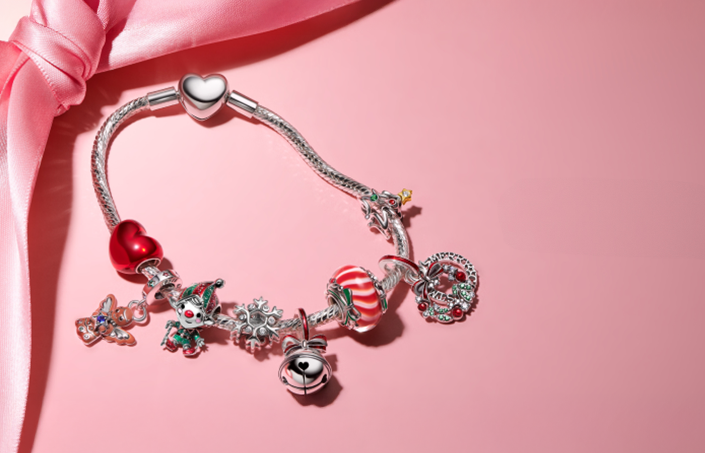 Pandora holiday charm bracelet featuring festive charms like a jingle bell, snowflake, candy cane, elf, and holiday wreath on a silver snake chain with heart clasp — styled with a pink ribbon on a soft pink background, perfect for seasonal gifting.